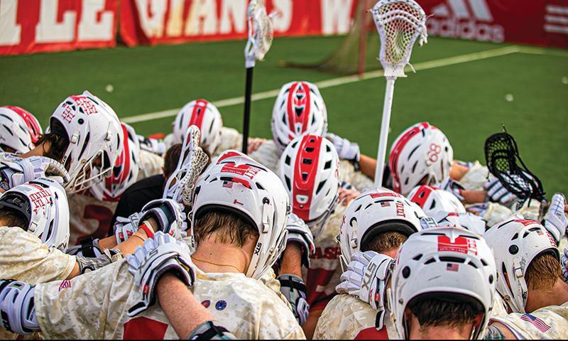A group of young men in lacrosse jerseys huddle with their backs to the camera